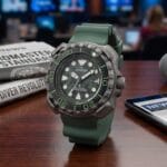 A Citizen Promaster watch with a green strap is displayed on a desk with a newspaper, tablet, and microphone in a news studio setting.