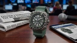 A Citizen Promaster watch with a green strap is displayed on a desk with a newspaper, tablet, and microphone in a news studio setting.