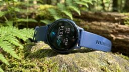A blue smartwatch displaying fitness tracking data rests on a mossy log in a forest setting with ferns in the background.