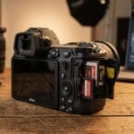 A digital camera with its dual SD card slots open sits on a wooden table near camera lenses and a laptop.