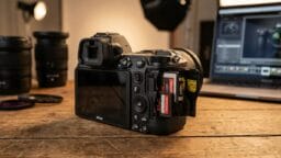 A digital camera with its dual SD card slots open sits on a wooden table near camera lenses and a laptop.