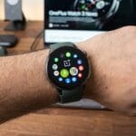A person wears a OnePlus smartwatch displaying various app icons on the home screen, with tech devices blurred in the background.