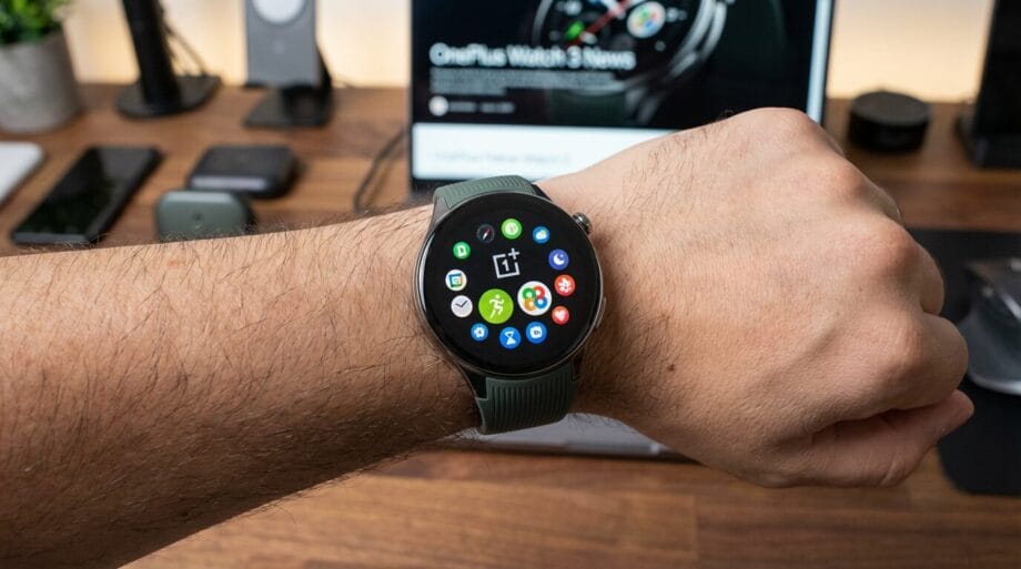 A person wears a OnePlus smartwatch displaying various app icons on the home screen, with tech devices blurred in the background.