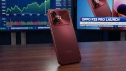A red Oppo smartphone stands on a wooden table with financial charts and an Oppo F33 Pro launch display in the background.