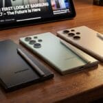 Three Samsung smartphones with styluses are displayed on a wooden surface, with a tablet showing a news article in the background.