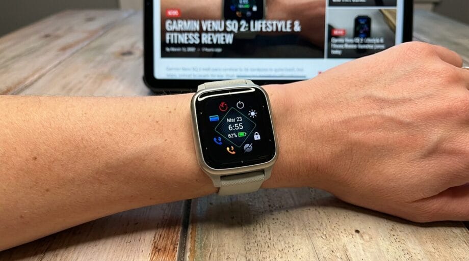 A person wearing a smartwatch with a digital display showing the time and date, with a tablet displaying a fitness review in the background.