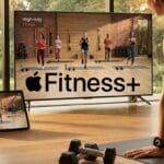 A person exercises indoors with Apple Fitness+ on a TV and tablet; dumbbells, a towel, and a water bottle are on the floor.