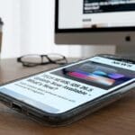 A smartphone displaying a news article about an iOS update rests on a desk with a coffee cup, glasses, and a computer monitor in the background.