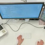 Person working at a desk with a laptop and two monitors displaying code, viewed from above.