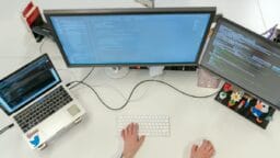 Person working at a desk with a laptop and two monitors displaying code, viewed from above.