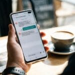 Person holding a smartphone displaying a text conversation, with a cup of coffee and a newspaper on the table in the background.