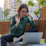 Person sitting on an outdoor bench, smiling and waving at a laptop during a video call, with buildings and trees in the background.