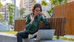 Person sitting on an outdoor bench, smiling and waving at a laptop during a video call, with buildings and trees in the background.
