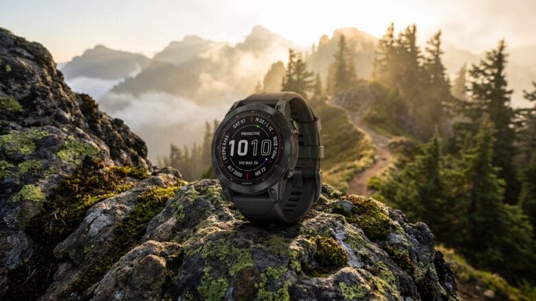 A black digital smartwatch is displayed on a mossy rock with a mountain landscape and pine trees in the background.