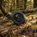 A digital outdoor watch rests on a mossy log in a sunlit forest, surrounded by fallen leaves and branches.