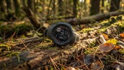 A digital outdoor watch rests on a mossy log in a sunlit forest, surrounded by fallen leaves and branches.