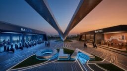 Futuristic Ola facility with electric scooters on the left, taxi service on the right, and illuminated paths converging at sunset.