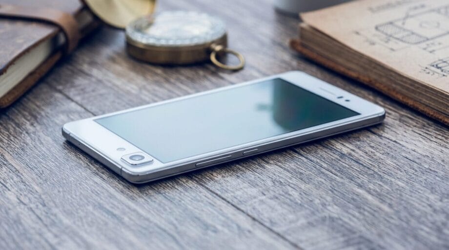 A silver smartphone lies face up on a wooden surface next to a notebook, a vintage compass, and a closed leather journal.