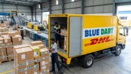 Workers load cardboard boxes into a Blue Dart DHL delivery truck inside a warehouse with conveyor belts and stacked parcels.