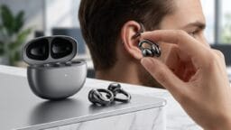 A person holds a sleek wireless earbud near their ear; an open charging case and two more earbuds rest on a counter.