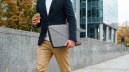 Person in business attire walks outside, holding a coffee cup and a gray laptop sleeve, with modern buildings in the background.