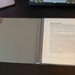 A digital notepad with a stylus is open on a desk, displaying a document. A laptop and keyboard are partially visible nearby.
