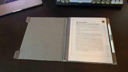 A digital notepad with a stylus is open on a desk, displaying a document. A laptop and keyboard are partially visible nearby.