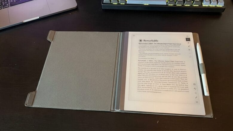 A digital notepad with a stylus is open on a desk, displaying a document. A laptop and keyboard are partially visible nearby.