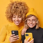 Two young women smiling and looking at smartphones against a solid yellow background.