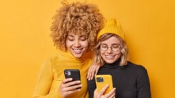 Two young women smiling and looking at smartphones against a solid yellow background.