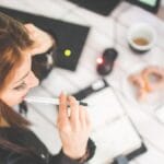 Woman sitting at a desk holding a pen, looking at an open planner, with a keyboard, coffee, and snacks nearby.