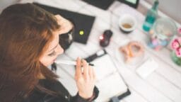 Woman sitting at a desk holding a pen, looking at an open planner, with a keyboard, coffee, and snacks nearby.