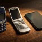 Three mobile phones on a wooden surface: a Nokia brick phone, a Motorola flip phone, and a modern touchscreen smartphone.