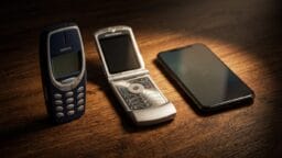 Three mobile phones on a wooden surface: a Nokia brick phone, a Motorola flip phone, and a modern touchscreen smartphone.